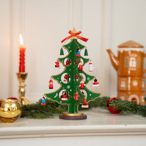 Small green wooden Christmas tree with miniature hanging ornaments shaped like Santa figures and red baubles, topped with a gold star and red ribbon, placed on a white surface next to a red candle, fir branches, and a gingerbread house-shaped teapot in the background.