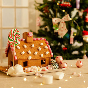 Gingerbread house decorated with small marshmallows, peppermint candies, and a red and green lollipop, placed on a tray surrounded by sweets and fairy lights, with a decorated Christmas tree in the background.