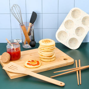 A stack of pancakes on a wooden cutting board with a jar of strawberry jam and various kitchen tools, including a whisk, spatula, and a speckled muffin tin.
