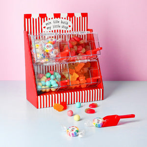 A toy candy dispenser display with a red and white striped design, filled with various candies like gumdrops, gummy bears, and jawbreakers, on a pink background.