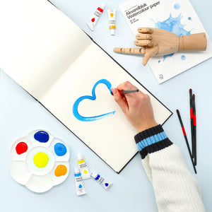Hand painting a blue heart with watercolours in a sketchbook, surrounded by paint tubes, brushes, palette, and a wooden mannequin hand.