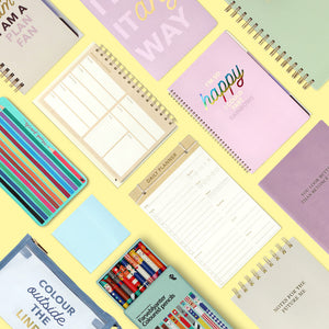A flat lay of colorful stationery and planners on a bright yellow background, including weekly and daily planners, spiral notebooks with inspirational quotes, and a set of colored pencils.
