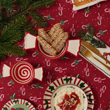 Festive Christmas table setting with red candy cane patterned tablecloth, striped plates, and candy-shaped bowls filled with cookies, surrounded by pine branches and gold cutlery wrapped in napkins.