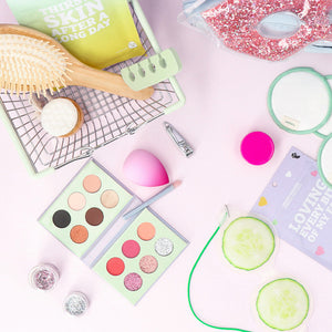 A flat lay of beauty and skincare products on a pink background, including an eyeshadow palette, makeup sponges, reusable cotton pads, a hairbrush, and a cucumber eye mask.