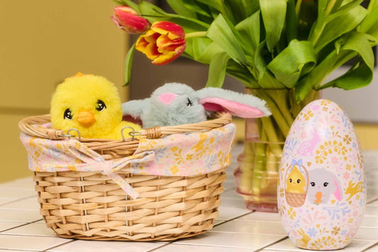 A basket with a plush soft toy chick and bunny, beside a decorated egg case, and flowers.