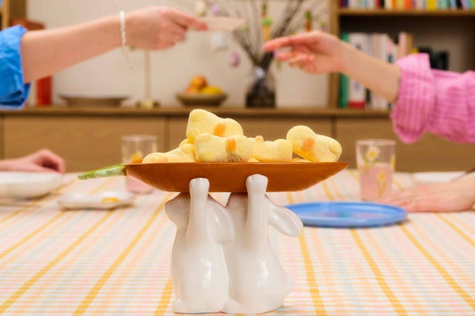 Easter get-together table centerpiece featuring a ceramic bunny bowl holder filled with festive marshmallow treats as guests share food in the background.