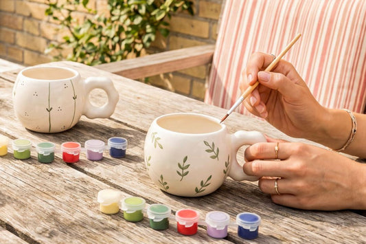 Person painting a ceramic mug with colorful paints on a wooden table.