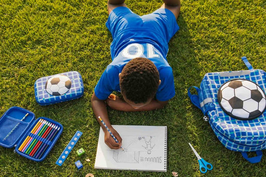 Child drawing on grass with art supplies and soccer-themed items nearby.