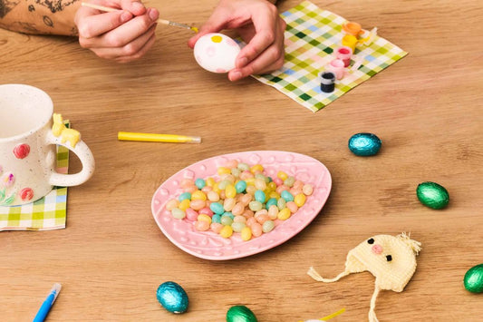 Hands painting an Easter egg with colorful candies and decorations on a table.