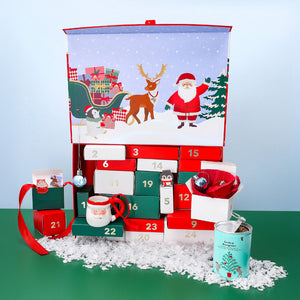 Colourful Christmas advent calendar box featuring Santa, a reindeer, and a sleigh illustration, with numbered drawers filled with small festive gifts, including a Santa mug, ornaments, and a tin of tea, displayed on faux snow.