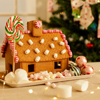 Gingerbread house on a Christmas tray with marshmallows, peppermint sweets, and a lollipop, lit by fairy lights with a Christmas tree in the background.