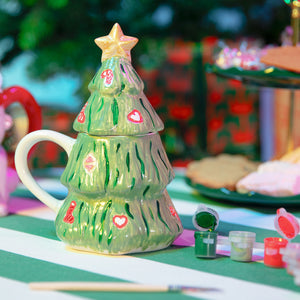 Festive ceramic mug shaped like a Christmas tree with a golden star on top, decorated with red hearts and candy cane designs. The mug sits on a green and white striped table beside small paint pots and a brush, with blurred Christmas cookies and wrapped presents in the background.