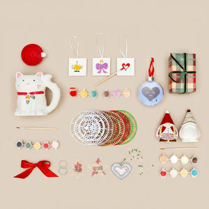 Flat lay of Christmas craft and décor items on a beige background, including a cat-shaped mug, paint sets, mini canvases with festive designs, gnome figurines, ornaments, ribbons, stencils, and a wrapped gift box.