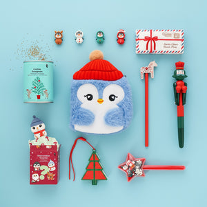 Flat lay of Christmas-themed gifts on a light blue background, including a plush penguin pouch with a red hat, festive pens, mini figurines, a tree-shaped ornament, a Santa letter box, and a small plant tin.