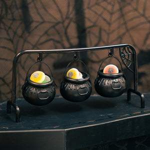 Three small black cauldrons hanging from a metal stand with a spiderweb detail, each filled with colourful gummy sweets, set against a dark Halloween-themed background with a web pattern.