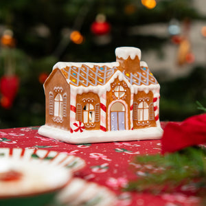Ceramic gingerbread house tealight holder on a red Christmas tablecloth, with blurred Christmas tree lights in the background.