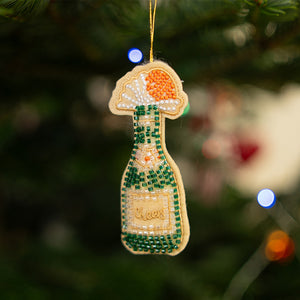 Beaded champagne bottle Christmas ornament with “Cheers” label hanging on a tree, blurred lights in the background.