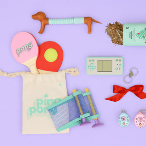 Flat lay of colourful gift ideas on a purple background, including ping pong paddles with a mesh net, a retro handheld game console, dog chew toy, keychains, red ribbon, mini plant tin, and small digital pets.
