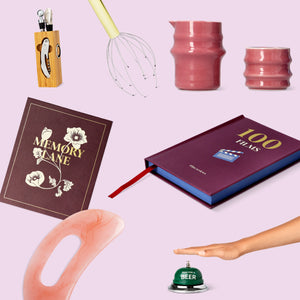 Flat lay of assorted gift items on a pastel pink background, including a book titled “100 Films,” a “Memory Lane” journal, pink ceramic cups, a gua sha tool, a beer service bell, a head massager, and a knife block set.