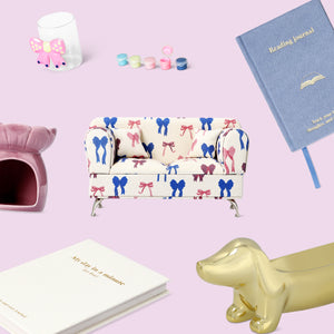 Flat lay of thoughtful gift items on a pastel pink background, including a patterned mini sofa with bow design, reading journal, “My day in a minute” notebook, pink pet house, gold dachshund figurine, and small pastel paint pots.