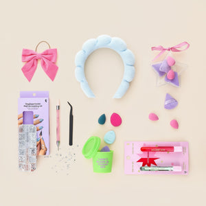 Flat lay of beauty and gift items on a beige background, including a blue padded headband, pink bow, makeup sponges, nail decorating kit, tweezer tool, glitter gems, and festive lip glosses in red and green packaging.