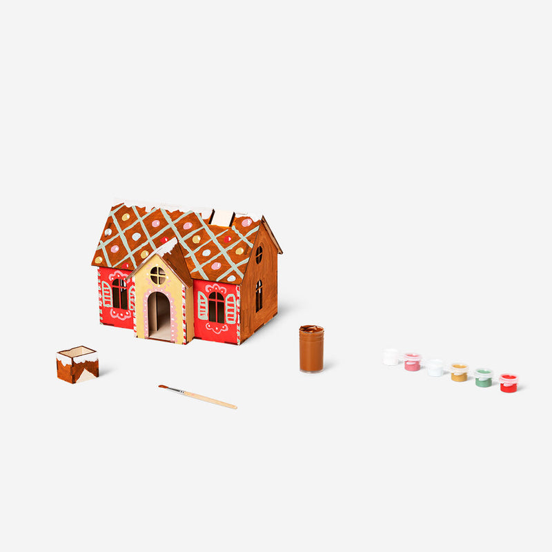 DIY gingerbread-style cardboard house craft set with painted details, surrounded by small paint pots, a paintbrush, and a jar of brown paint, displayed on a plain white background.