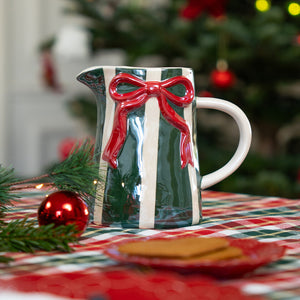 Ceramic jug with a glossy green and white design featuring a raised red bow on the front, placed on a red and green checkered tablecloth with Christmas decorations and a plate of cookies in the foreground, and a decorated Christmas tree in the background.
