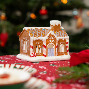 Ceramic gingerbread house with candy cane pillars and white icing details, placed on a red festive tablecloth with Christmas patterns and blurred decorations in the background.