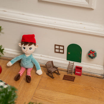 Plush elf toy with red hat and striped leggings beside a miniature green door scene, tiny ladder, rocking chair, welcome mat, red post box, window and wreath on a wall.